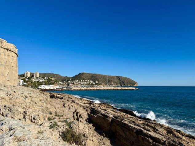 Вид на прибрежный ландшафт Теулада-Морайра вдоль геологического маршрута, с скальными образованиями на переднем плане и замком и портом Морайра на заднем плане.