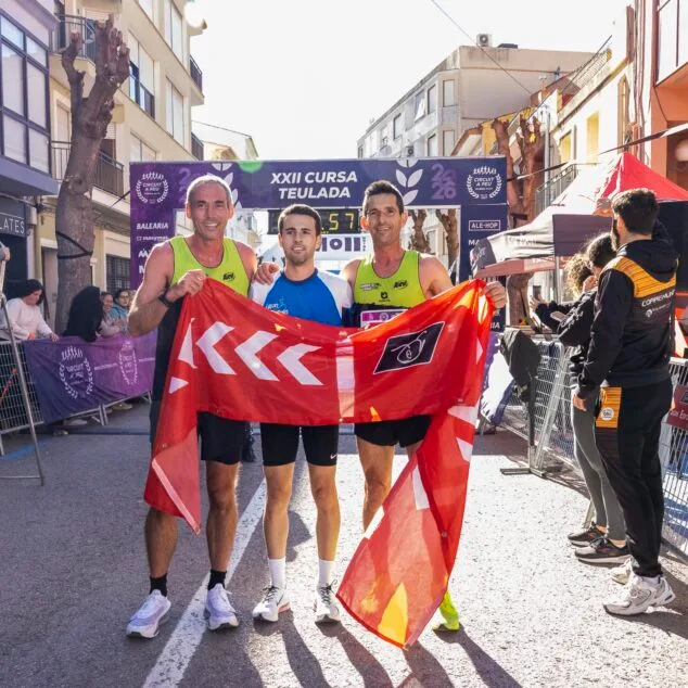 Imagen: Tres corredores masculinos ganadores posan con una bandera en la meta de la XXII Volta Popular Teulada.
