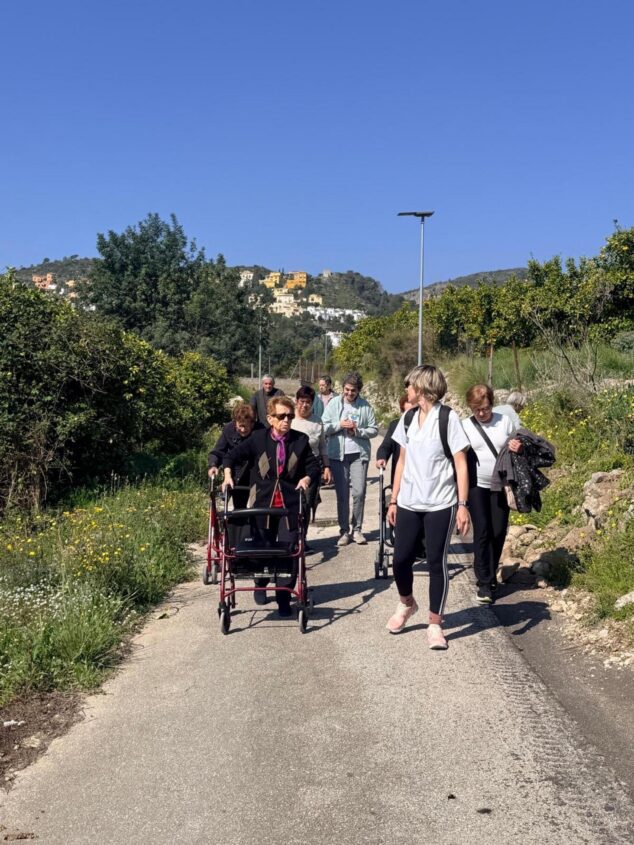 Imagen: Grupo de personas mayores, una de ellas con andador, caminando por una ruta rural acompañadas por una profesional sanitaria.