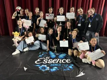 Un grupo de jóvenes bailarinas en un escenario sosteniendo diplomas y trofeos después de una competición.