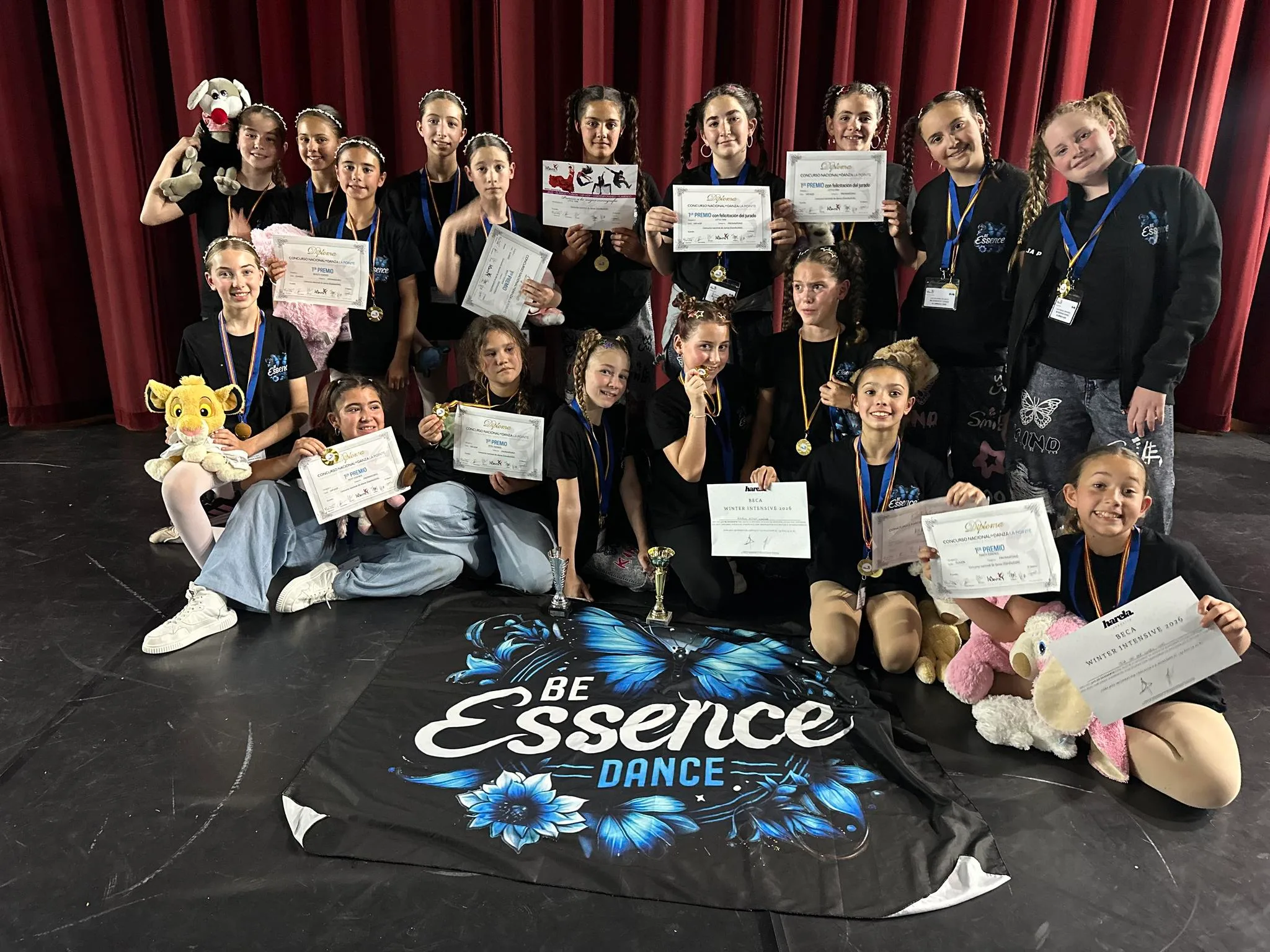 Un grupo de jóvenes bailarinas en un escenario sosteniendo diplomas y trofeos después de una competición.