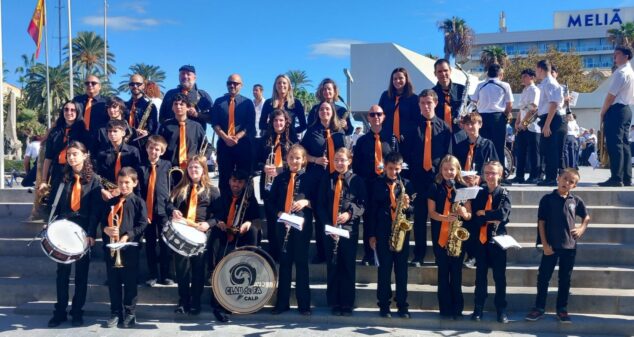 Foto de grupo de los miembros de la Agrupació Musical CLAUdeFA–CALP con sus instrumentos y uniforme negro con corbata naranja en unas escaleras exteriores.