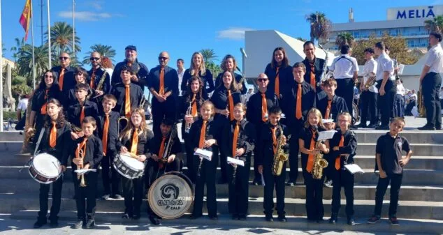 Imatge: Foto de grup dels membres de l'Agrupació Musical CLAUdeFA–CALP amb els seus instruments i uniforme negre amb corbata taronja a unes escales exteriors.