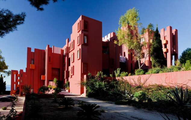 Imagen: Vista exterior del edificio Muralla Roja de Ricardo Bofill en Calp, con su característica arquitectura geométrica de color rojo y rosa entre vegetación.