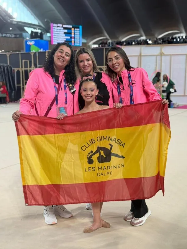 Yulen Rubio, la promesa de Calp que conquista España en gimnasia 1 Imagen: Gimnasta y entrenadoras del CG Les Marines Calpe sostienen una bandera de España con el logo del club en un pabellón deportivo.