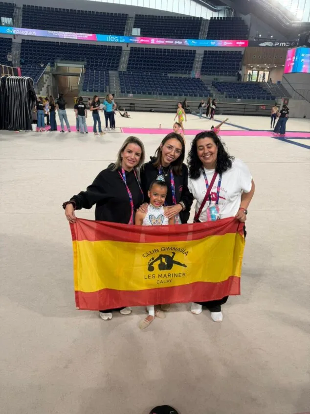 Yulen Rubio, la promesa de Calp que conquista España en gimnasia 6 Imagen: Una joven gimnasta y tres mujeres del equipo técnico del Club Gimnasia Les Marines de Calp sostienen una bandera española con el logo del club dentro de un gran pabellón deportivo.