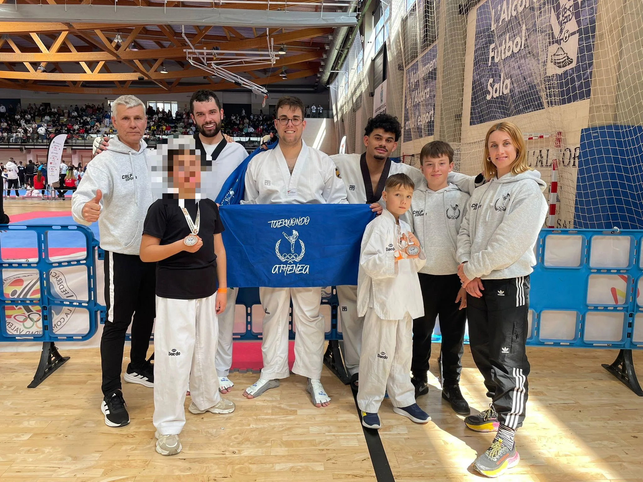 Integrantes del Club de Taekwondo Athenea de Calp posan en grupo con una bandera y medallas en un pabellón deportivo.
