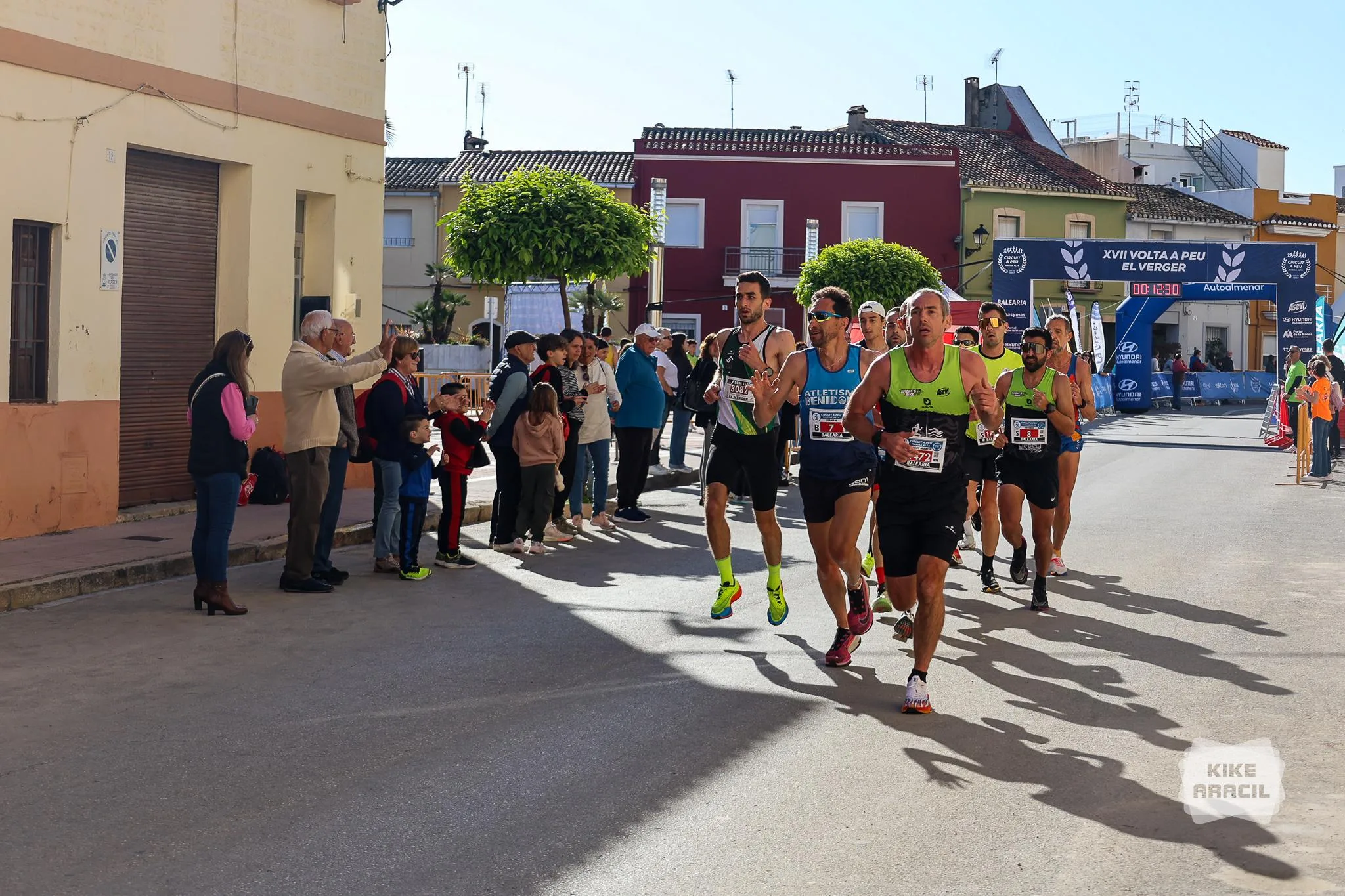 Varios corredores disputan la carrera Volta a Peu El Verger, con espectadores animando y un arco de meta al fondo.