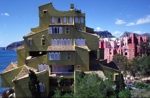 Imagen: Fotografía exterior del singular edificio Xanadú de color amarillo verdoso, con tejados de teja y contraventanas blancas, situado en un acantilado junto al mar en Calp.
