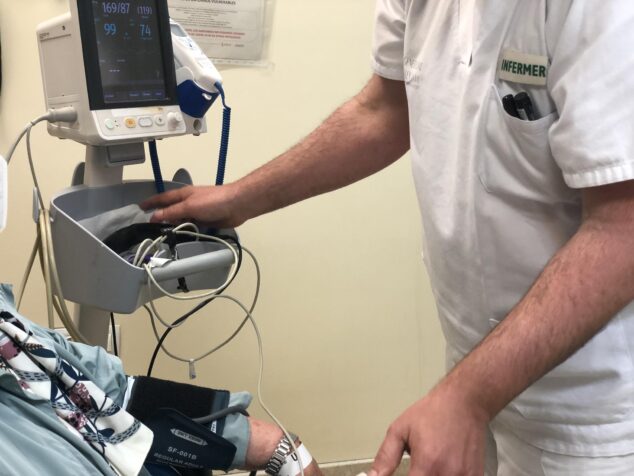Imagen: Un enfermero con uniforme blanco y el texto "INFERMER" toma la presión arterial a un paciente, cuyo brazo tiene un manguito conectado a un monitor de signos vitales.