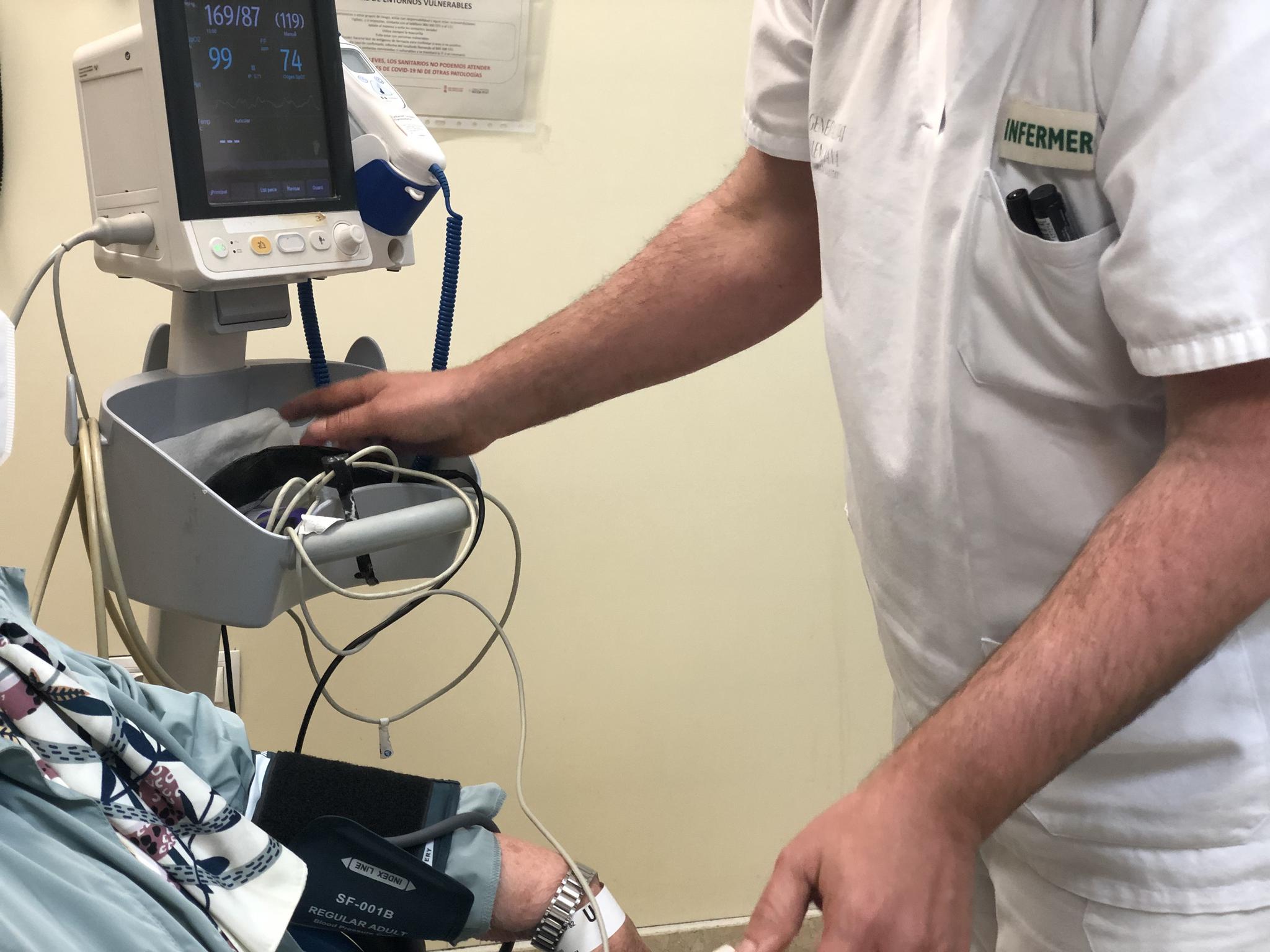 Un enfermero con uniforme blanco y el texto "INFERMER" toma la presión arterial a un paciente, cuyo brazo tiene un manguito conectado a un monitor de signos vitales.
