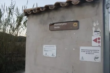 Fachada de hormigón del punto de información de la Muntanyeta Verda con un cartel marrón que indica el nombre y varios paneles informativos blancos sobre proyectos de adecuación en la Marjal de Pego-Oliva.