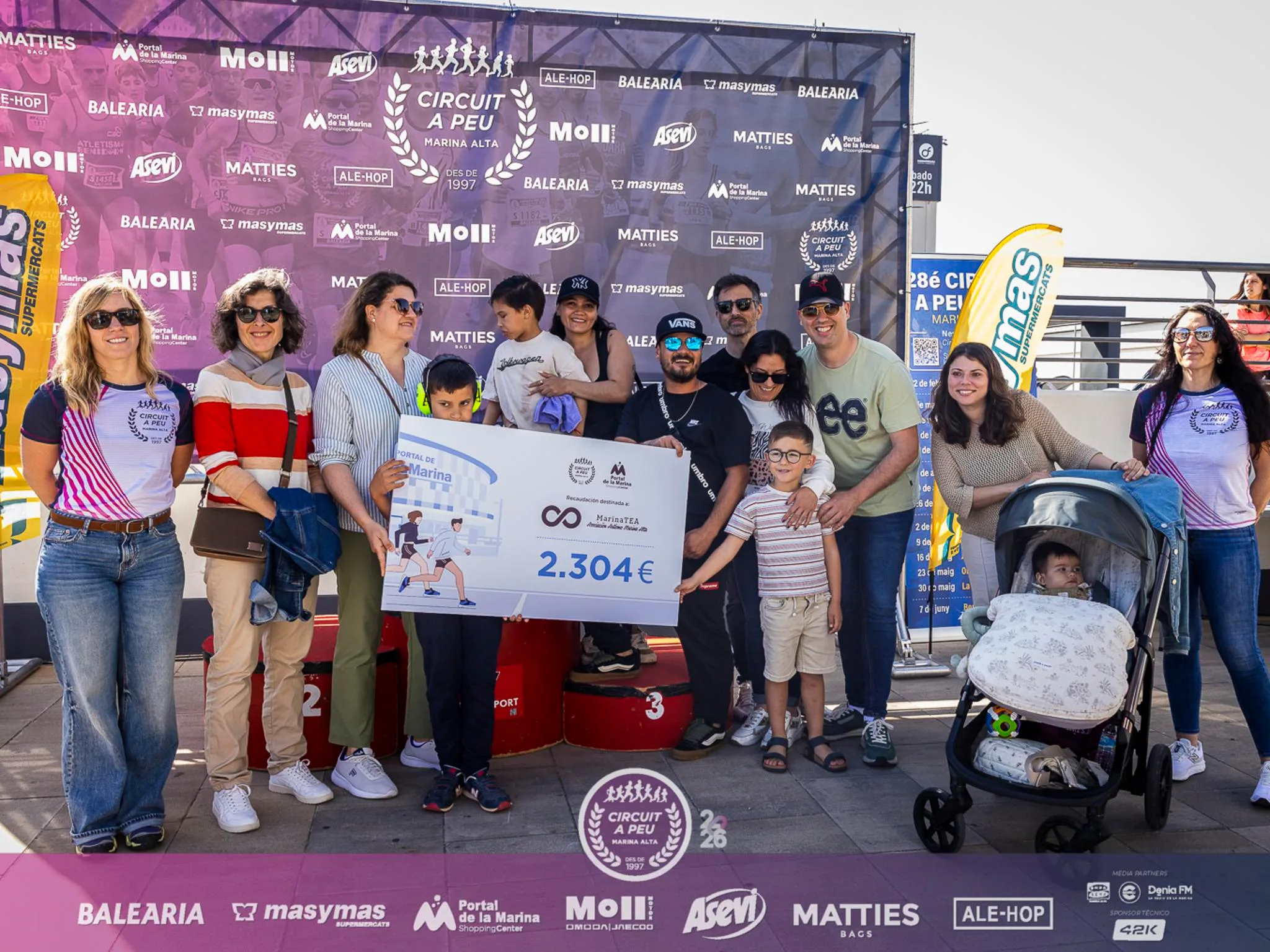 Foto de grupo con un cheque gigante de 2.304 euros para la asociación MarinaTEA frente al photocall del Circuit a Peu Marina Alta.