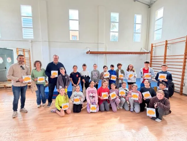 Grupo de escolares con libros y diplomas posando junto a dos adultos en un gimnasio escolar durante una entrega de premios.