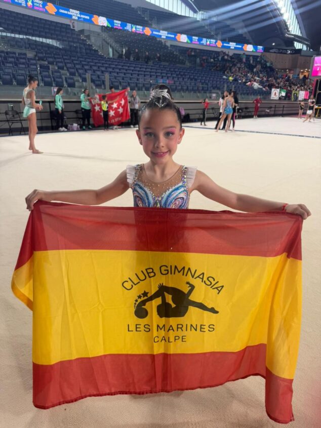 Imagen: Una joven gimnasta del Club Les Marines de Calp sonríe sosteniendo una bandera de España con el logo del club en un pabellón deportivo.