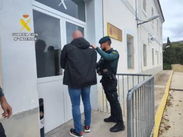 Un agente de la Guardia Civil escolta a un hombre detenido, con la cara pixelada, hacia la entrada de un edificio oficial.