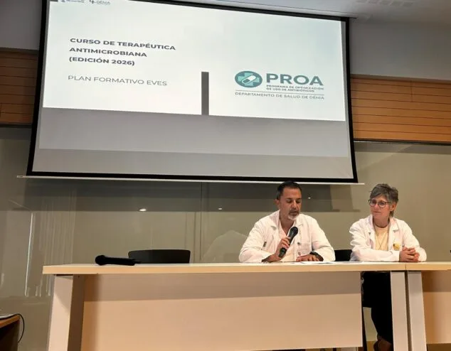Imagen: Juan Puig Larrosa, gerente del Departamento de Salud de Dénia, habla con un micrófono en una mesa junto a otra profesional sanitaria, frente a una pantalla que anuncia el "Curso de Terapéutica Antimicrobiana (Edición 2026)" y el programa PROA.