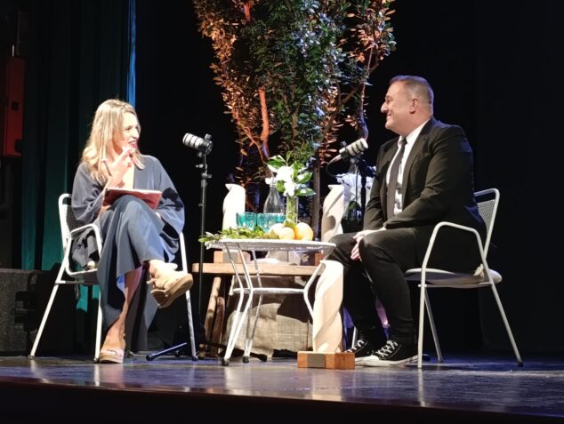 Imagen: El escritor Jovi Lozano-Seser, a la derecha, sentado en un escenario durante la gala de los Premios Vila de Santanyí, conversando con una presentadora.