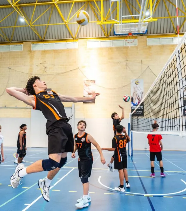 Imagen: Jugador de voleibol con camiseta negra y naranja número 89 saltando para rematar el balón por encima de la red durante un partido en un pabellón cubierto, con otros jugadores observando.