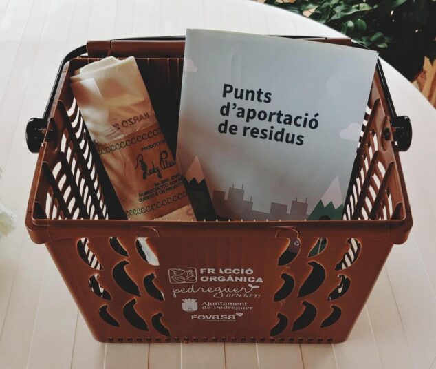 Cesta de recogida de orgánica con el logo del Ajuntament de Pedreguer y folleto sobre puntos de aportación de residuos.