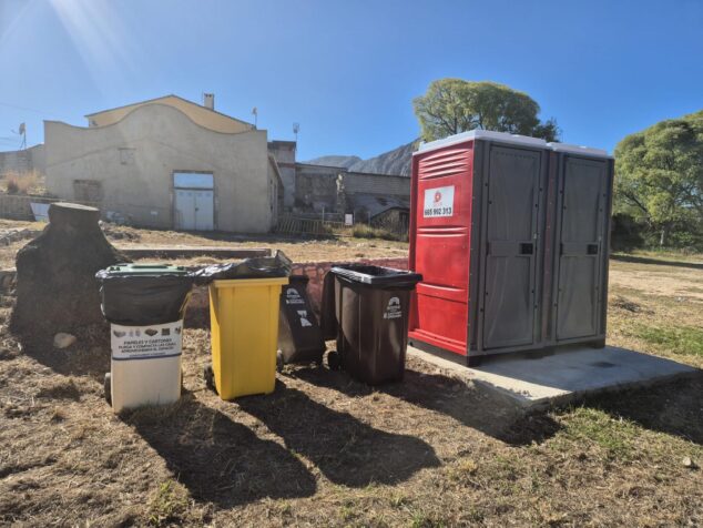 Imagen: Aseos portátiles y contenedores de reciclaje en el área recreativa del Molí Coví en Ondara, con un edificio antiguo al fondo.