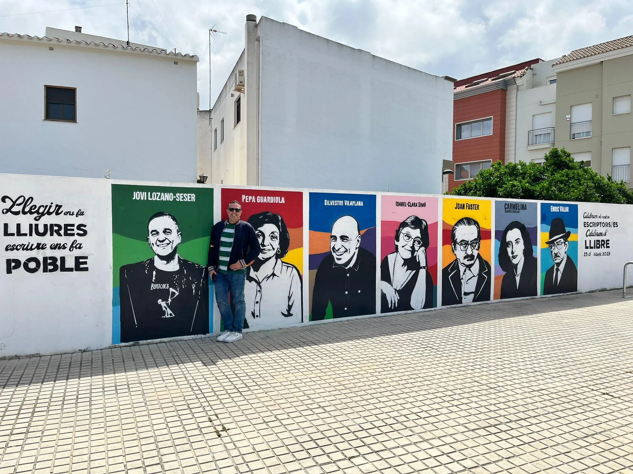 Jovi Lozano-Seser de pie junto a su retrato en un colorido mural al aire libre dedicado a escritores valencianos en Ondara.