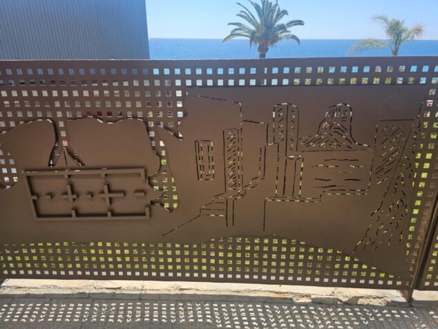 Imagen: Primer plano de la nueva barandilla artística de metal en Calp, con siluetas recortadas de edificios y vistas al mar y palmeras de fondo