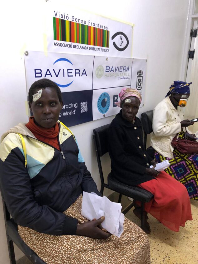 Tres mujeres africanas sentadas con vendajes en los ojos en una sala de espera, con carteles de Visió Sense Fronteres y Baviera al fondo.