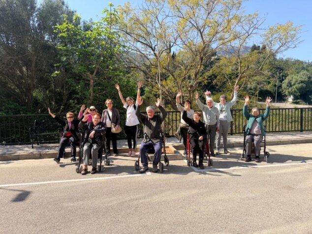 Imagen: Grupo de personas mayores y adultos saludando, algunos sentados en andadores, al aire libre durante la actividad saludable "Camina".