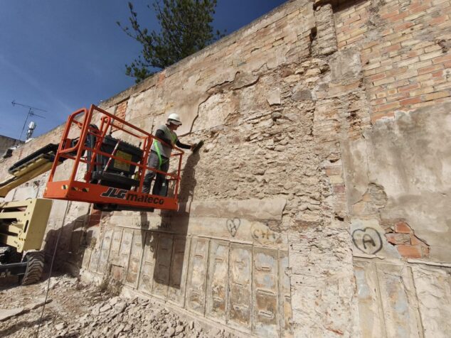 Un trabajador en una plataforma elevadora naranja inspecciona el antiguo muro de piedra y ladrillo del Trapig del Verger durante trabajos arqueológicos.