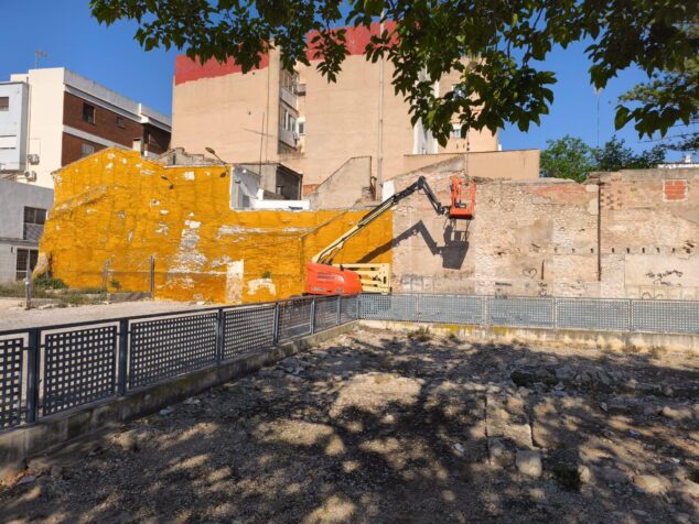 Imagen: Una excavadora amarilla con plataforma trabaja junto a un muro antiguo de piedra y ladrillo en un yacimiento arqueológico.