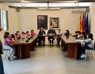 El alcalde Sergi Ferrús y la concejala Noelia Garcia sentados en una mesa grande con un grupo de niños pequeños en el salón de plenos del Ayuntamiento de Pedreguer.