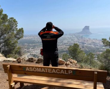 El vehículo rápido de Protección Civil de Calp interviene en 21 incendios 117 Un miembro de Protección Civil de Calp, de espaldas, mira con prismáticos desde un mirador con un banco que dice #ASÓMATEACALP y vistas al Peñón de Ifach.