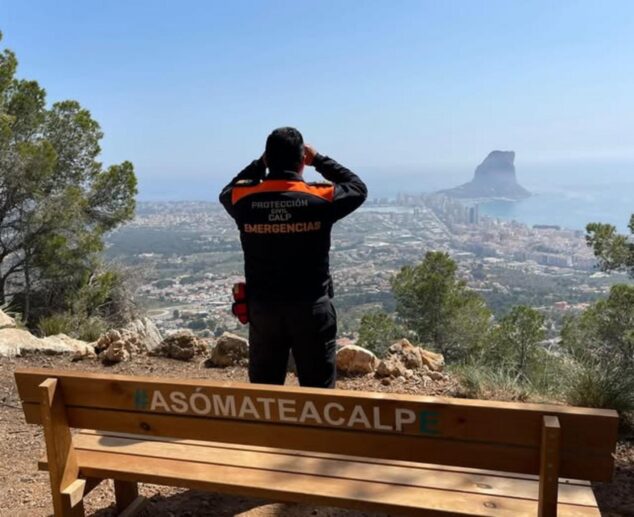 Imagen: Un miembro de Protección Civil de Calp, de espaldas, mira con prismáticos desde un mirador con un banco que dice #ASÓMATEACALP y vistas al Peñón de Ifach.