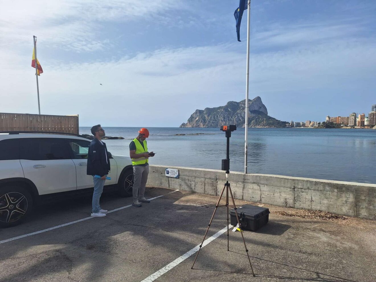 Dos técnicos con equipo de topografía trabajan junto al mar en Calp, con el Peñón de Ifach al fondo.