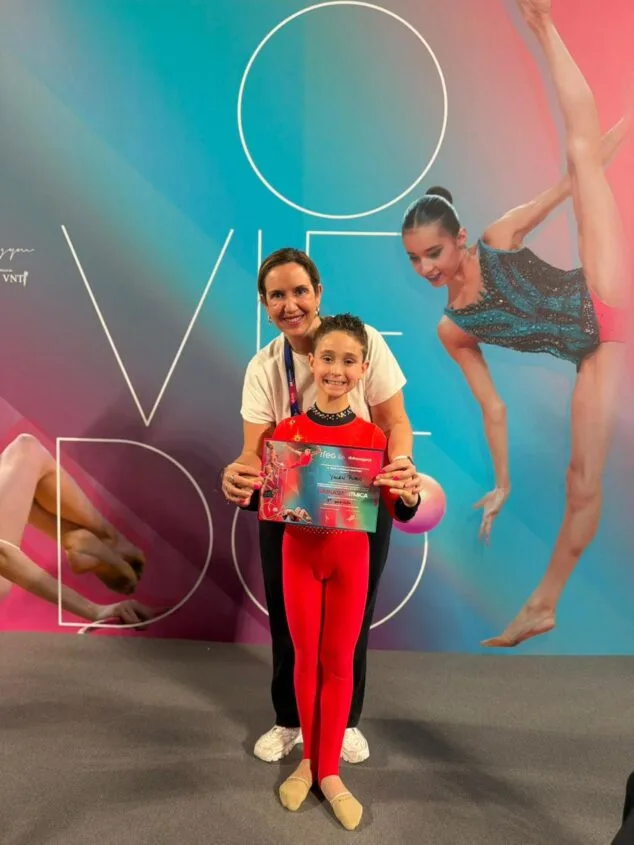 Yulen Rubio, la promesa de Calp que conquista España en gimnasia 3 Imagen: Yulen Rubio, joven gimnasta masculino, sostiene un diploma junto a su entrenadora frente a un cartel de campeonato.