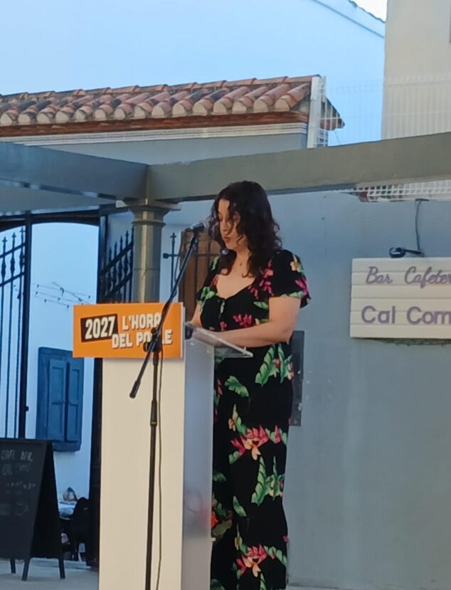 Compromís conmemora 15 años en la alcaldía de Pedreguer y activa su precampaña para 2027 9 Una mujer habla desde un atril con el lema "2027 L'Hora del Poble" en un acto de Compromís al aire libre.