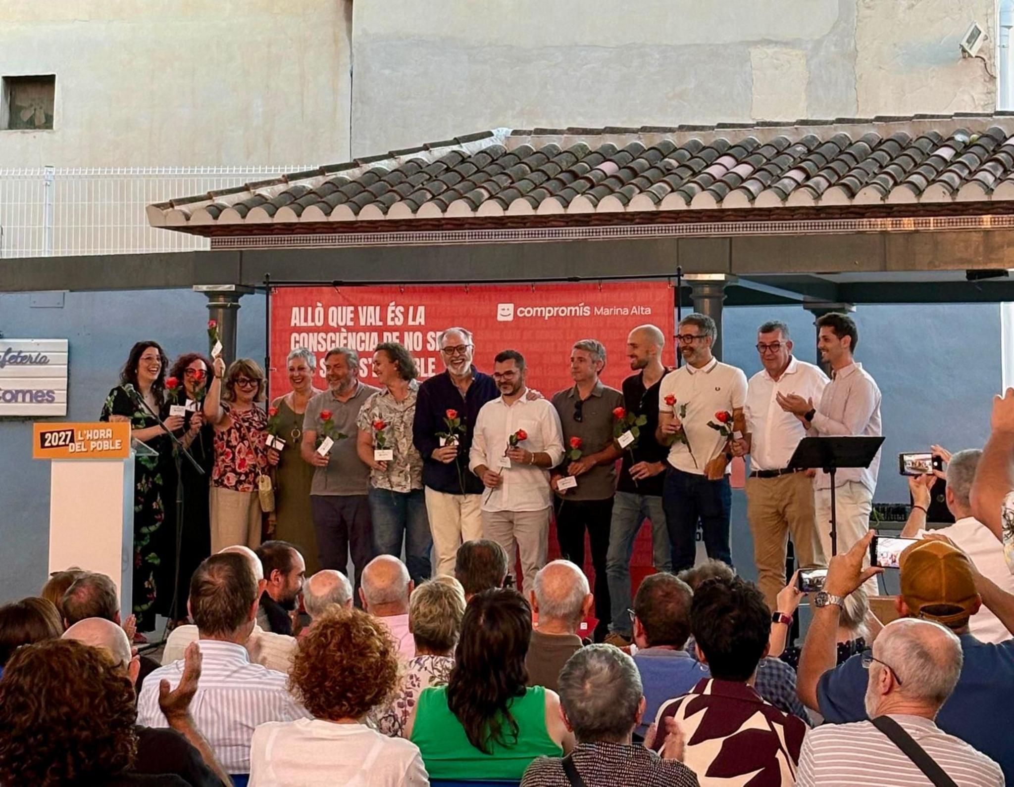 Grupo de representantes políticos de Compromís sosteniendo rosas en un escenario al aire libre ante el público.