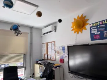 Ondara invierte en mejorar la climatización del CEIP Sanchis Guarner 16 Vista interior de un aula escolar con una unidad de aire acondicionado tipo split montada en la pared blanca sobre un corcho de anuncios.