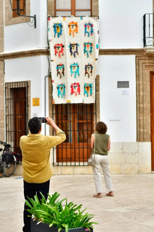 Imagen: Gente mirando una gran obra de arte textil con patrones colgada de un balcón en una plaza del pueblo.