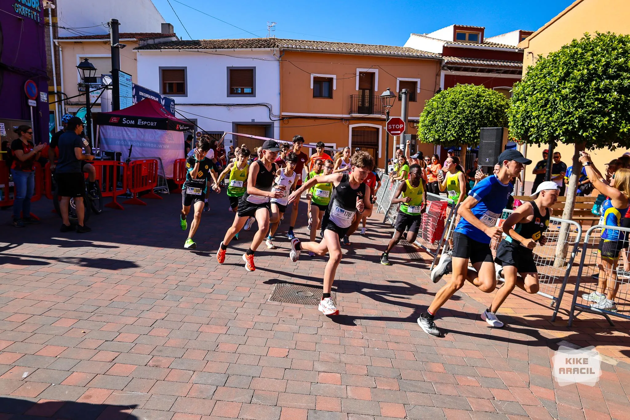 Varios niños y niñas corren tras la salida en una carrera urbana con público y un arco de meta.