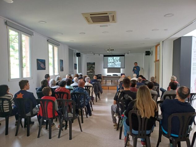 Imagen: Un conferenciante habla con micrófono ante un público sentado en una sala iluminada con una pantalla de proyección al fondo.