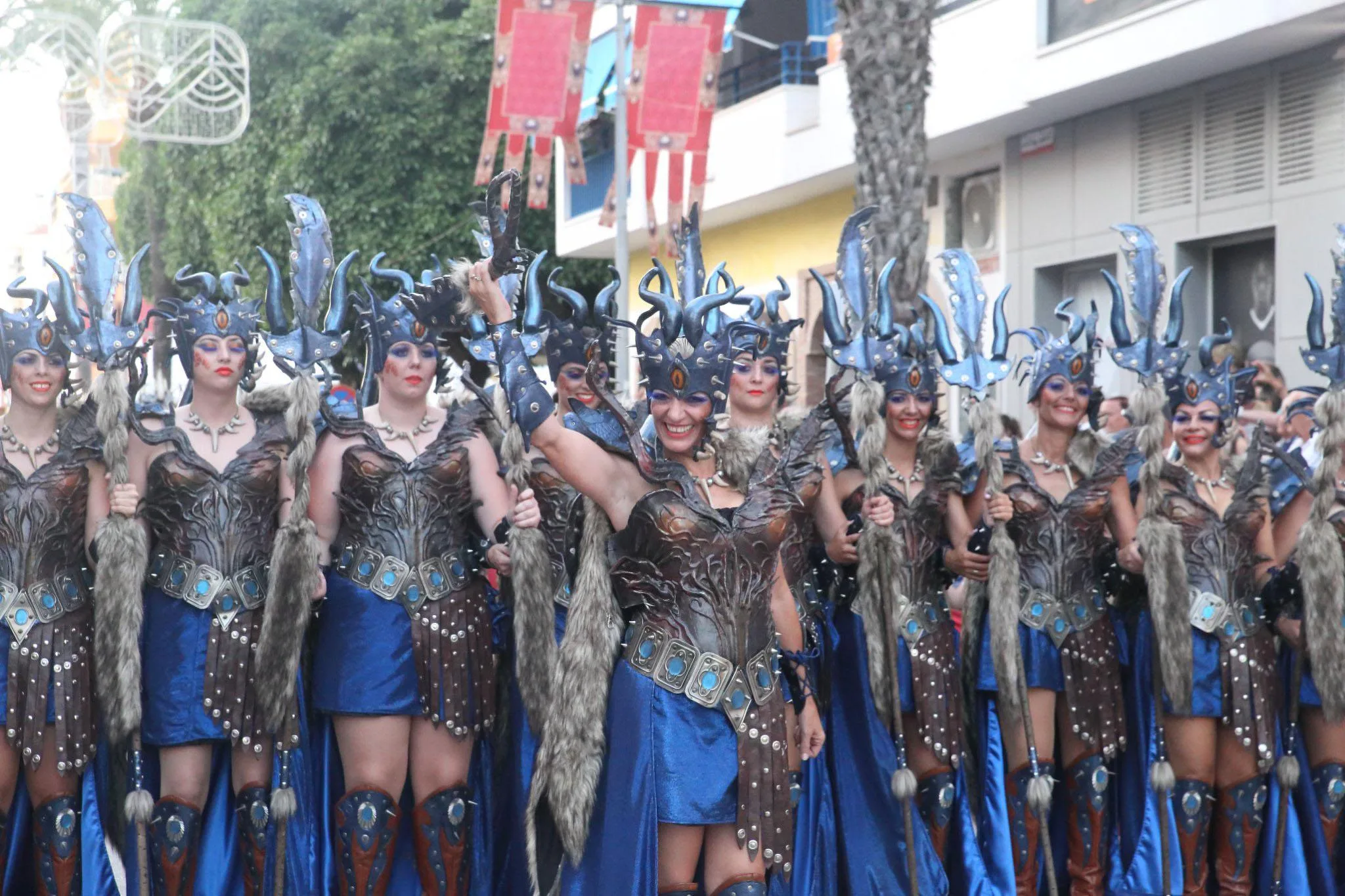 Un grupo de mujeres con trajes de guerreras desfilan en una calle decorada con estandartes durante una fiesta.
