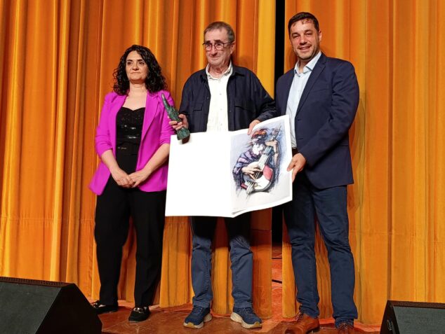 Rosa Ana Marí y José Ramiro entregan el premio Ocell y una ilustración al profesor Josep Puig Ramis en un escenario con cortinas naranjas.