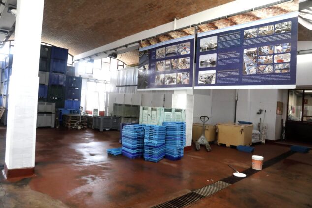 Interior de una lonja de pescado con pilas de cajas de plástico azules y grises y paneles informativos en la pared de fondo.