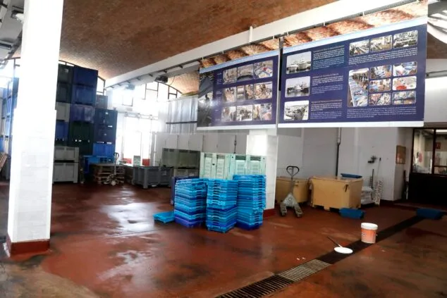 Image : Intérieur d'un marché aux poissons avec des piles de caisses en plastique bleu et gris et des panneaux d'information sur le mur du fond.