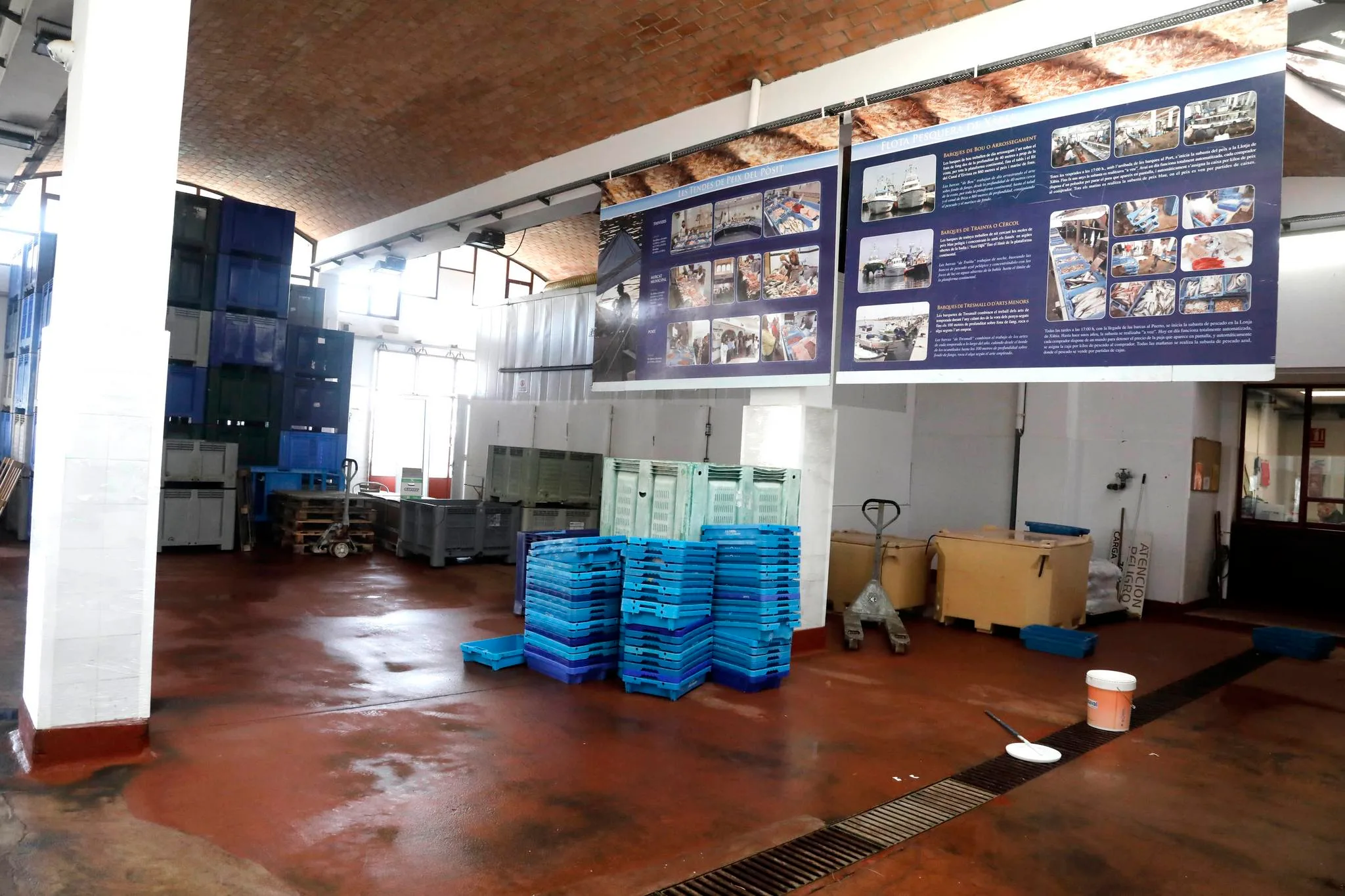 Interior de una lonja de pescado con pilas de cajas de plástico azules y grises y paneles informativos en la pared de fondo.