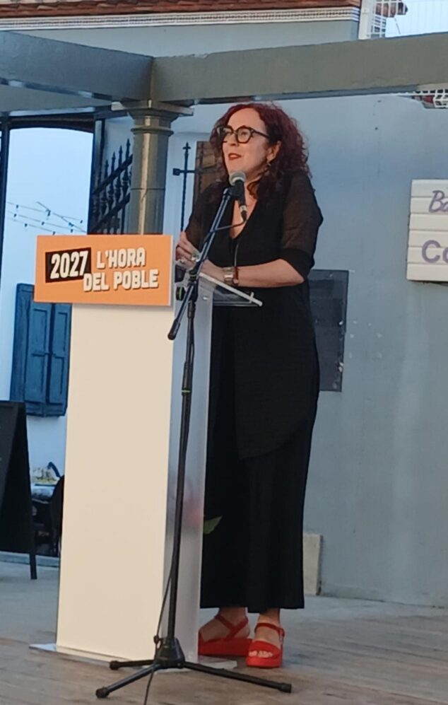 Compromís conmemora 15 años en la alcaldía de Pedreguer y activa su precampaña para 2027 1 Imagen: Mujer hablando en un atril con el lema '2027 L'Hora del Poble' durante un acto político al aire libre.