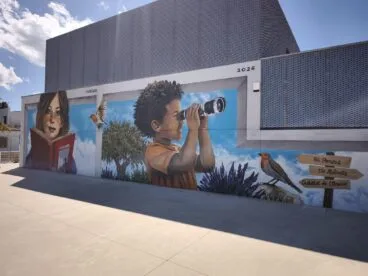 El CEIP L'Alfàs de Pedreguer estrena un mural de Tardor con referencias locales 24 Fotografía del gran mural en la pared del CEIP L'Alfàs en Pedreguer, mostrando a niños aprendiendo y elementos del paisaje local como señales hacia el Castell de l'Ocaive.