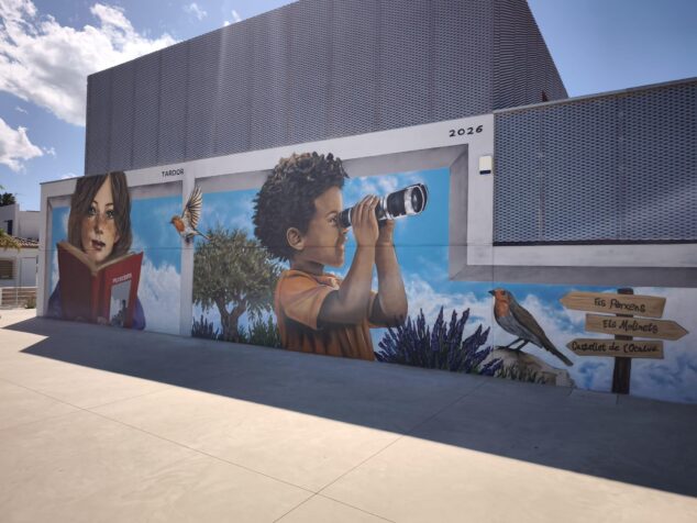 Imagen: Fotografía del gran mural en la pared del CEIP L'Alfàs en Pedreguer, mostrando a niños aprendiendo y elementos del paisaje local como señales hacia el Castell de l'Ocaive.